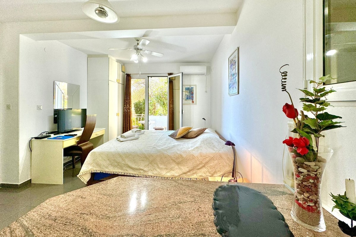A modern bedroom with a bed, a desk, a chair, a television, and a window with a view of greenery.