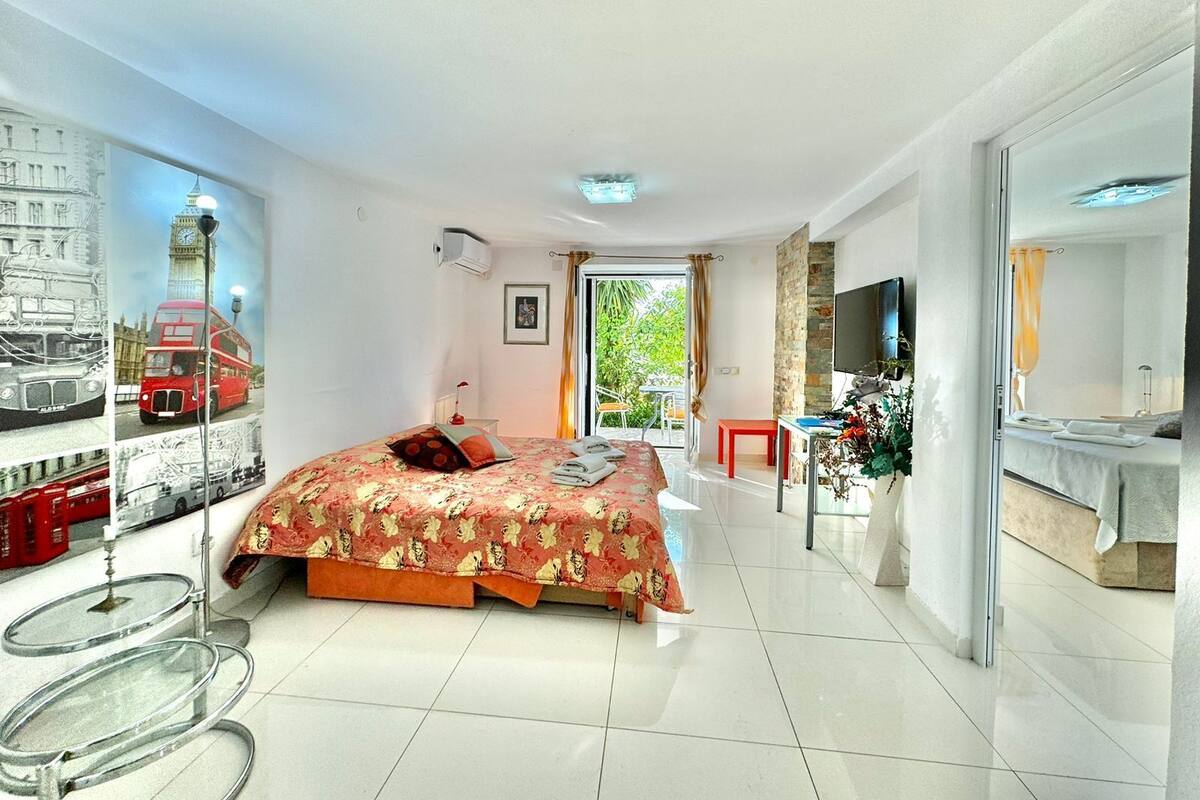 A modern hotel room with a bed, a TV, a bathroom, and a balcony with a view of greenery.