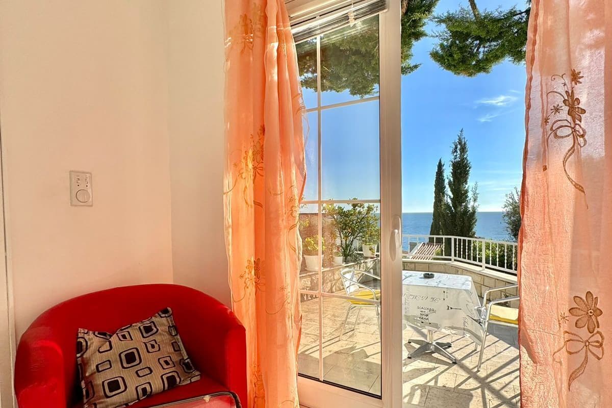 A room with a red chair, a small table, and a view of the sea through a glass door.