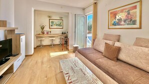 A modern living room with a sofa, a coffee table, and a TV. There is a kitchen area with a dining table and chairs. - Sun Rose apartments Sveti Stefan (Sveti Stefan)