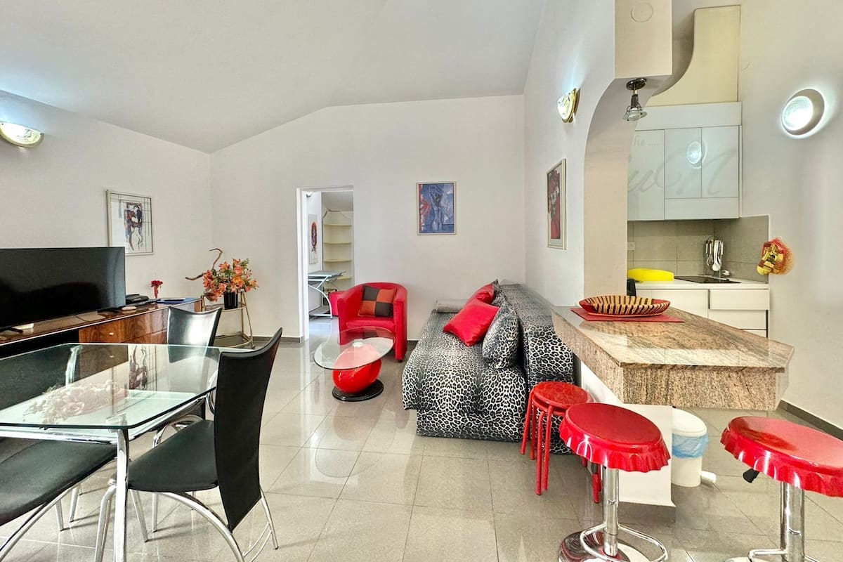 A modern living room with a glass dining table, red chairs, a sofa with leopard print cushions, and a kitchen area with a sink and cabinets.
