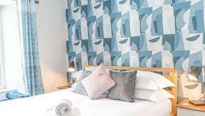 Una cama bien hecha con toallas enrolladas, un papel tapiz estampado y una mesita de noche con lámpara.