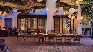 Un área de comedor al aire libre con mesas y sillas, rodeada de árboles y faroles colgantes.