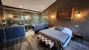 Un dormitorio moderno con una cama grande, un banco, una mesita pequeña y un baño visible a través de una mampara de cristal.