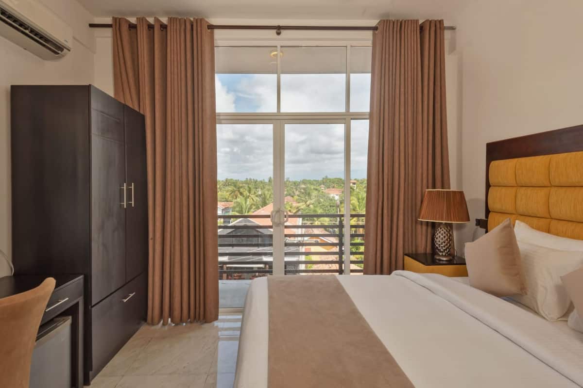 A hotel room with a bed, a wardrobe, a nightstand with a lamp, and a view of the outdoors.