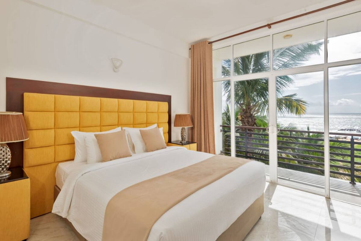 A hotel room with a large bed, a nightstand, a lamp, and a view of the ocean through large windows.