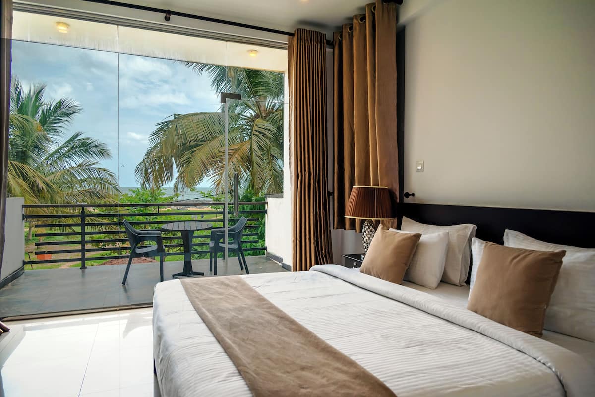 A hotel room with a large bed, a balcony with a table and chairs, and a view of palm trees and the ocean.