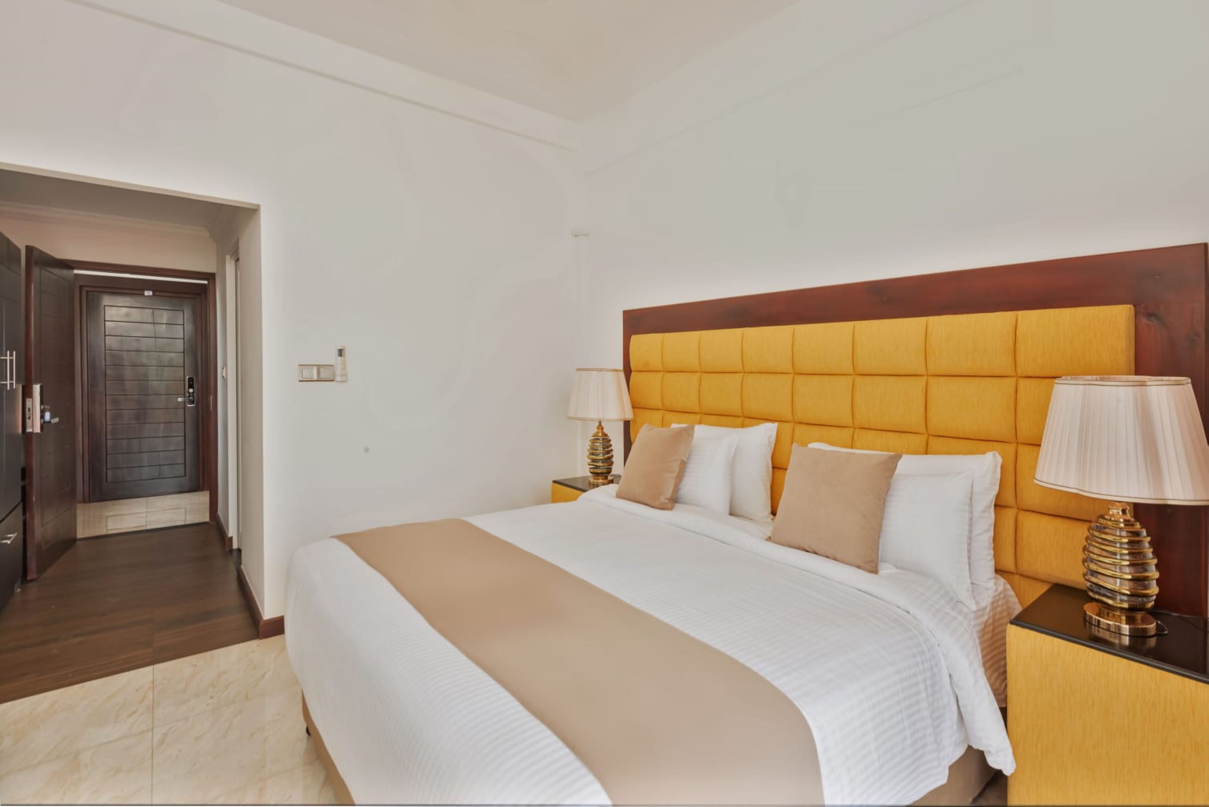 Quarto de hotel com uma cama grande, duas mesas de cabeceira, uma cabeceira de madeira e uma porta que dá para um corredor.