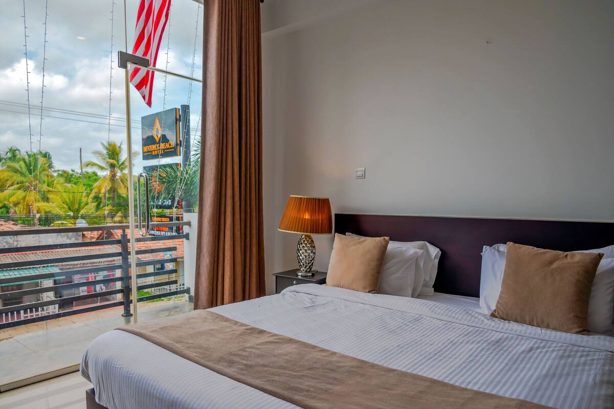 A hotel room with a bed, a nightstand, a lamp, and a view of the outside.