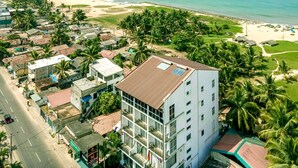 A multi-story hotel with a flat roof and large windows, located near a beach with palm trees and a road. - Beverly Beach - Negambo (Negombo)