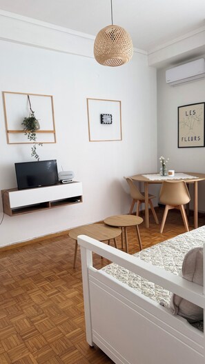 A modern, minimalist room with a white bed, a wooden floor, a TV on a white console, a wicker pendant light, a small round table with chairs, and framed pictures on the wall.