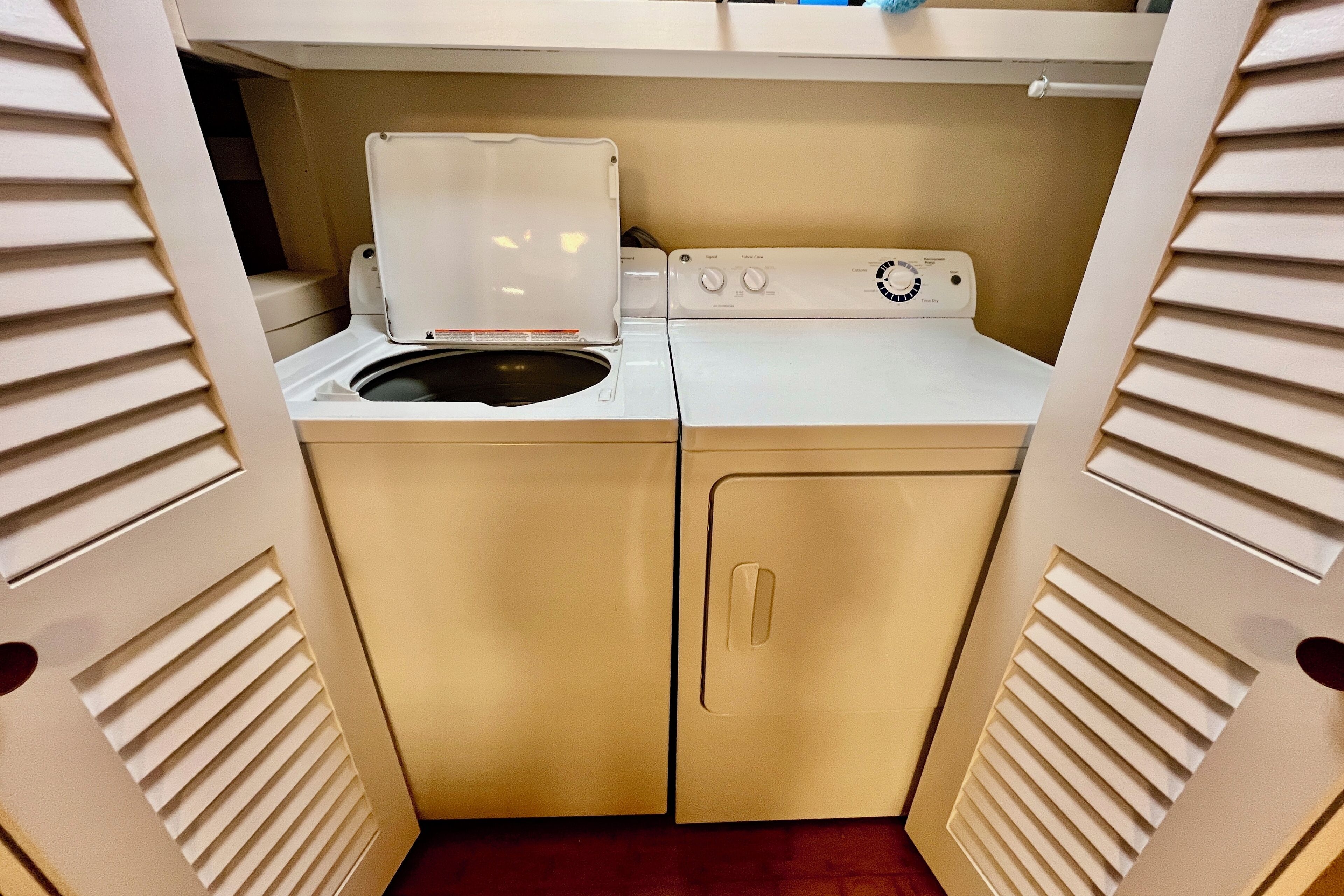 Une petite buanderie comprenant un lave-linge et un sèche-linge, le tout intégré dans un espace délimité par des murs de couleur beige et doté de grilles de ventilation.