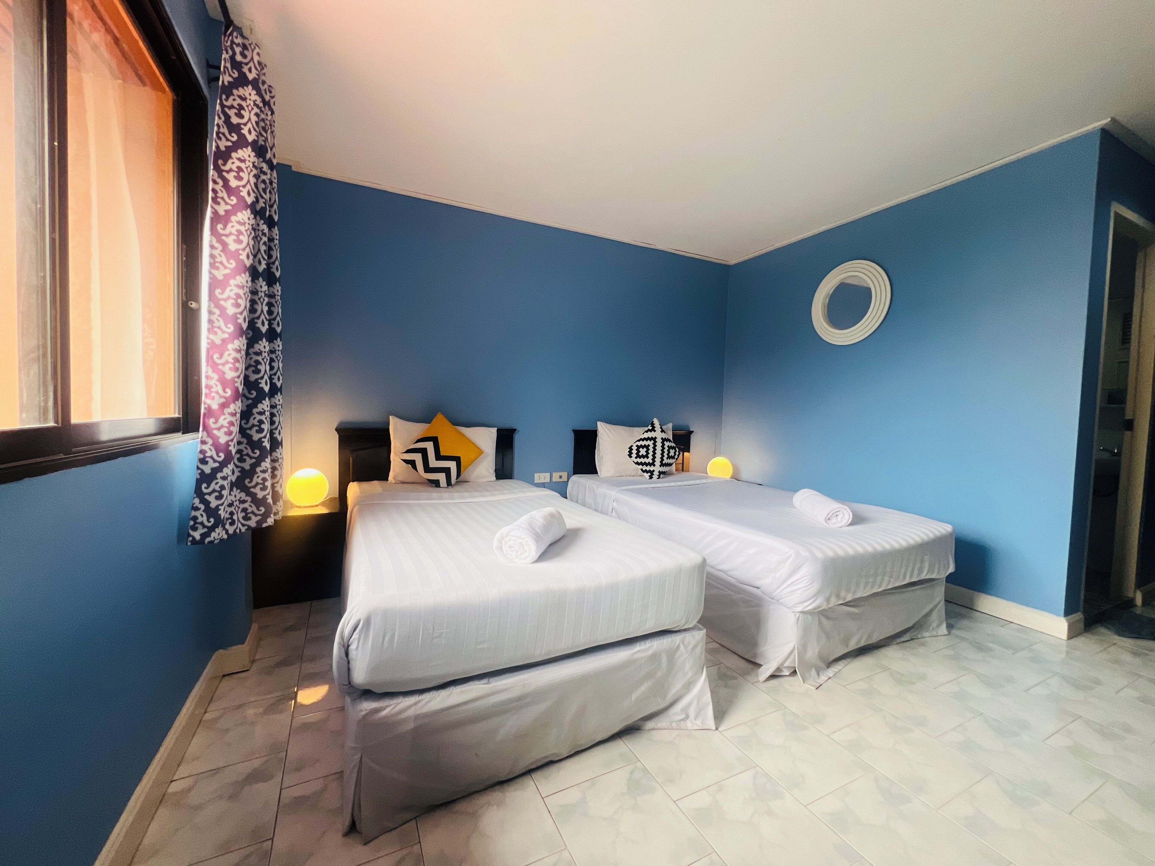 Una habitación con dos camas, una ventana con cortinas estampadas, un adorno circular en la pared y suelo de baldosas.