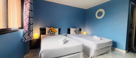 Una habitación con dos camas, una ventana con cortinas estampadas, un adorno circular en la pared y suelo de baldosas.