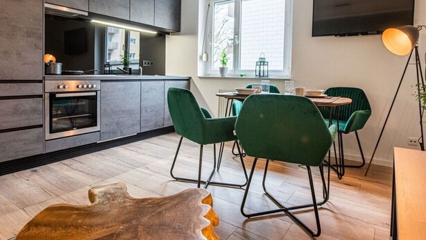 Une cuisine moderne avec des meubles de couleur sombre, un four encastré et un coin repas agrémenté de chaises vertes et d’une table ronde.