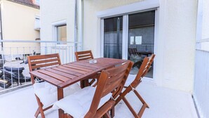 Una mesa y sillas de madera en un balcón con barandal blanco, una puerta corrediza de vidrio y vistas a un edificio cercano.