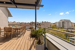 Eine Holzterrasse mit Sitzgelegenheiten im Freien und einem Blumentopf, mit Blick auf eine Stadtlandschaft und Gebäude.
