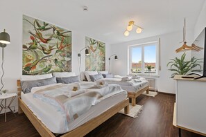 Ein modernes Hotelzimmer mit drei Betten, großen abstrakten Gemälden, einem Schreibtisch mit Lampe und Blick ins Freie.