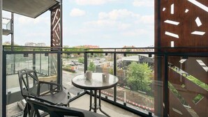 Un balcón con mesa y sillas, ventanas amplias que dan a edificios y una calle, y un panel de madera decorativo con un reloj.