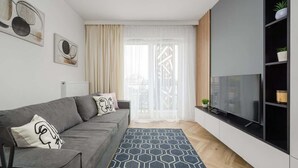 Una sala de estar moderna con un sofá gris, un televisor de pantalla plana grande, una alfombra estampada y una ventana con cortinas traslúcidas.
