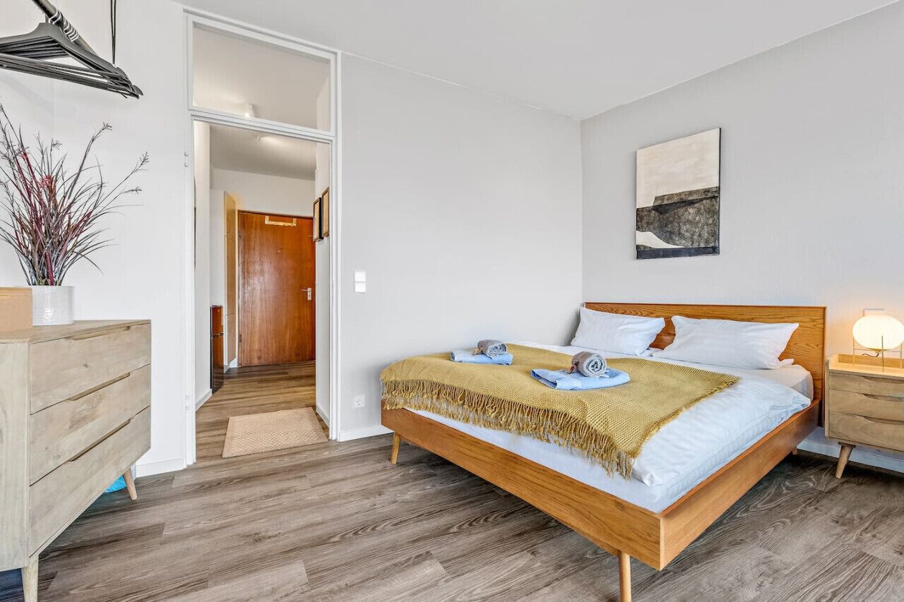 Quarto com cama de madeira, mesa de cabeceira com abajur, porta que dá para outro cômodo e planta em vaso.