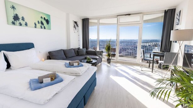 Una camera d'albergo moderna con un letto grande, una poltrona, un'area ristoro e un balcone con vista sulla città.