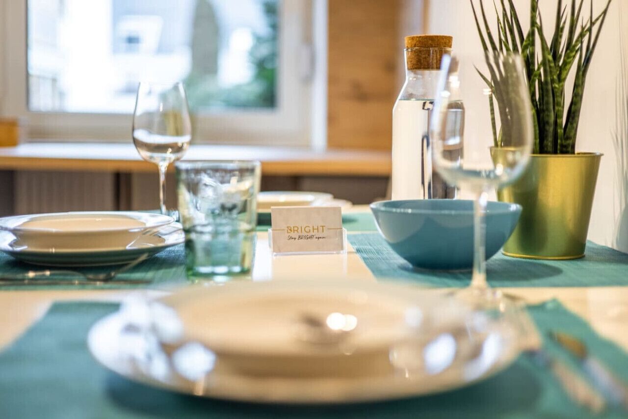 Une table dressée avec un verre transparent, une assiette blanche, un bol bleu, une petite carte, une bouteille et une plante en pot.