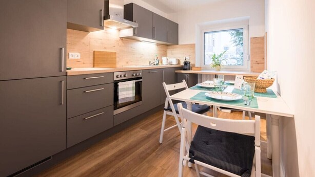 A modern kitchen with grey cabinets, a wooden countertop, and a dining area with white chairs and a table set for two. - Arbio I Beach style Apart (Augsburg)