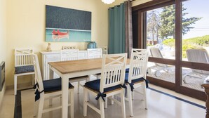 Uma sala de jantar com uma mesa de madeira e cadeiras brancas, um quadro na parede e vista através de uma porta de vidro.