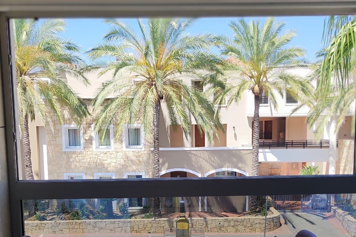 A dining area with a table and two chairs, a bottle of wine, and a view of palm trees and buildings through a window.