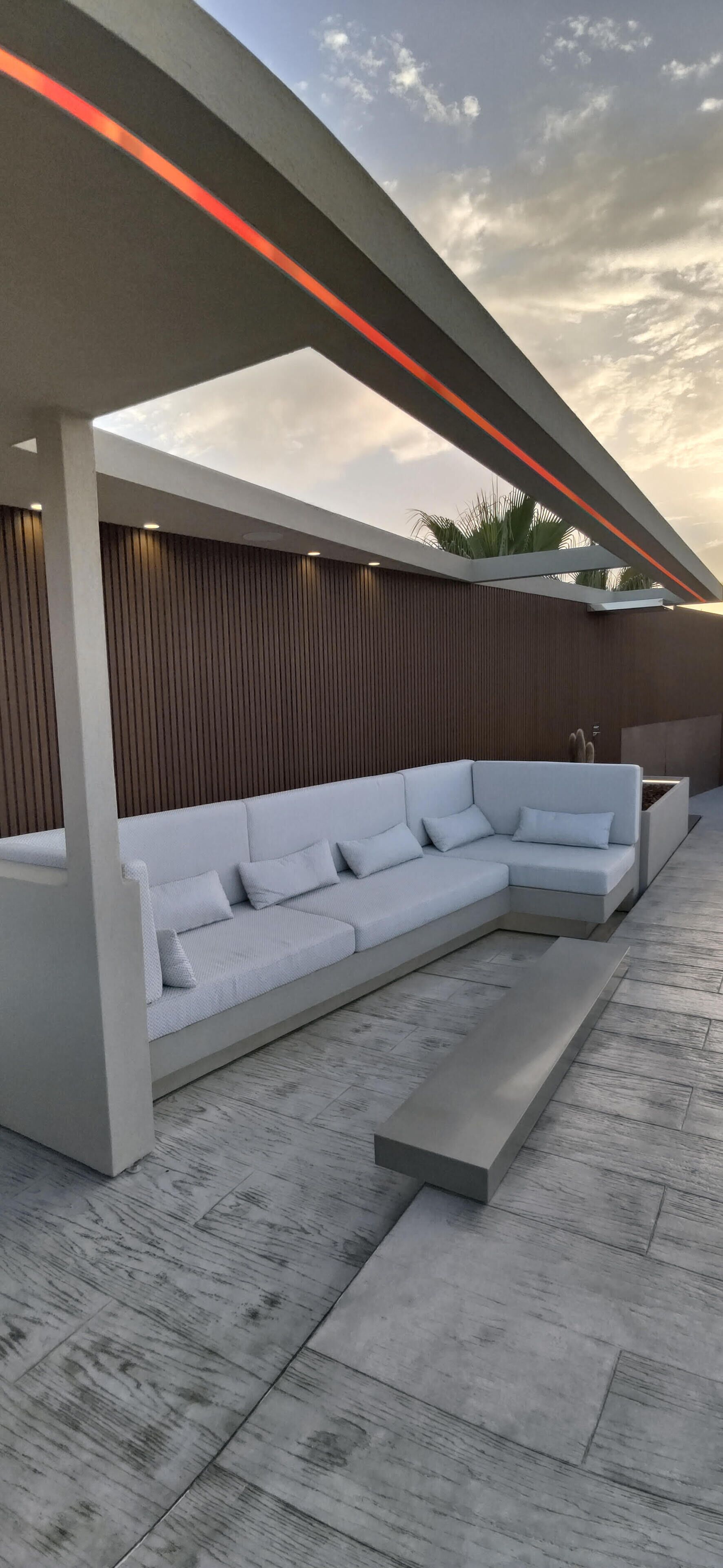 A modern outdoor seating area with a white sectional sofa, a wooden slatted wall, and a concrete floor.
