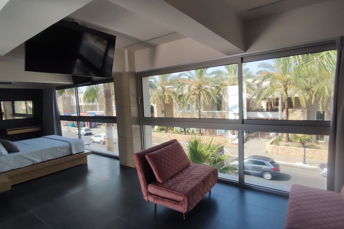 A modern hotel room with a large window, a bed, a sofa, and a TV mounted on the ceiling.