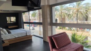 Una camera da letto moderna con un letto, una poltrona rosa, una TV e ampie finestre con vista su palme.