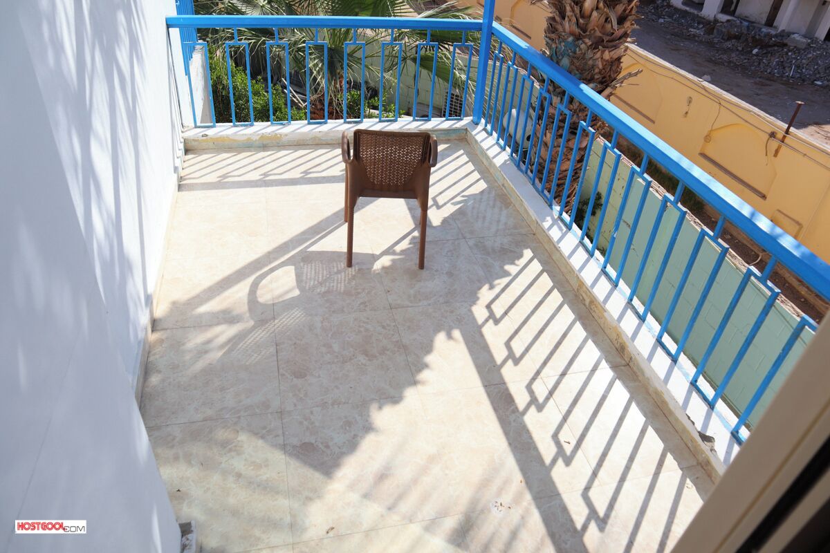 A balcony with a chair, blue railing, and a view of a building and palm trees.