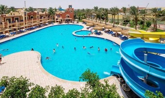 A large swimming pool with a water slide, surrounded by lounge chairs and palm trees.