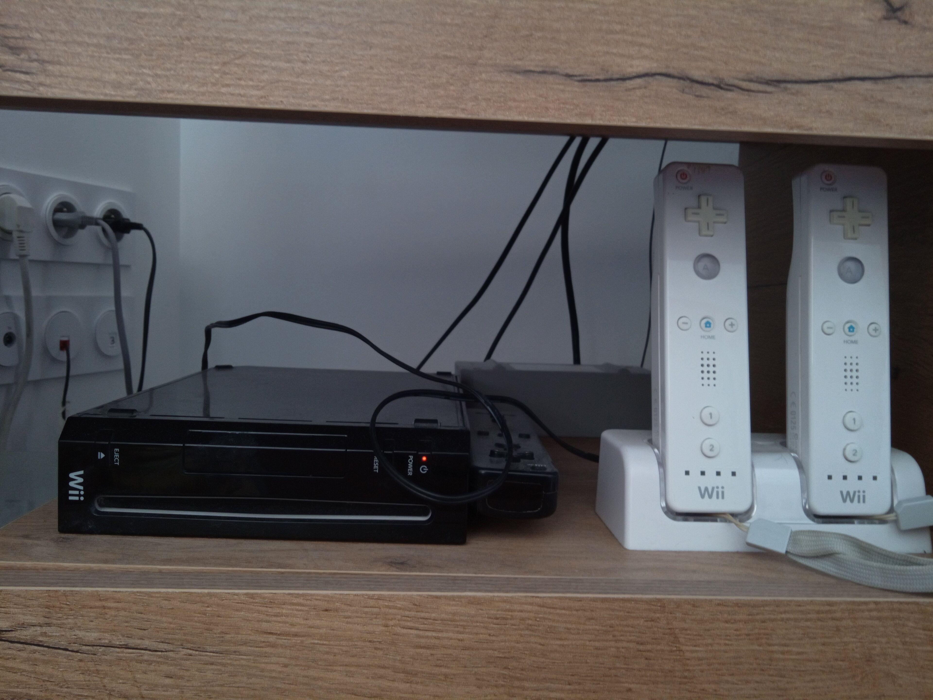 En svart Wii-konsol är ansluten till två vita Wii-kontroller som står på en hylla av trä.