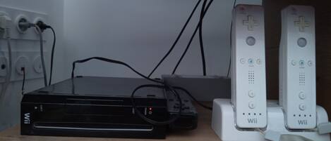 En svart Wii-konsol är ansluten till två vita Wii-kontroller som står på en hylla av trä.