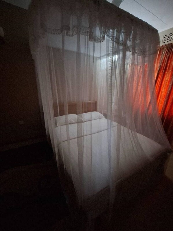 Habitació amb un llit cobert per un teulat prim, una finestra visible amb cortines i una vista parcial d'una paret.