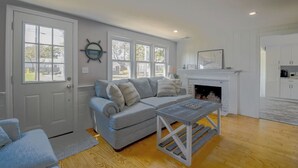 A living room with a gray sofa, a fireplace, and a kitchen area visible through an open door. - Beachy 3 bedroom home with central air, just .5 miles to the beach (Dennis Port)