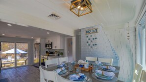 A dining area with a table set for six, white chairs, and a kitchen in the background. - Beachy 3 bedroom home with central air, just .5 miles to the beach (Dennis Port)