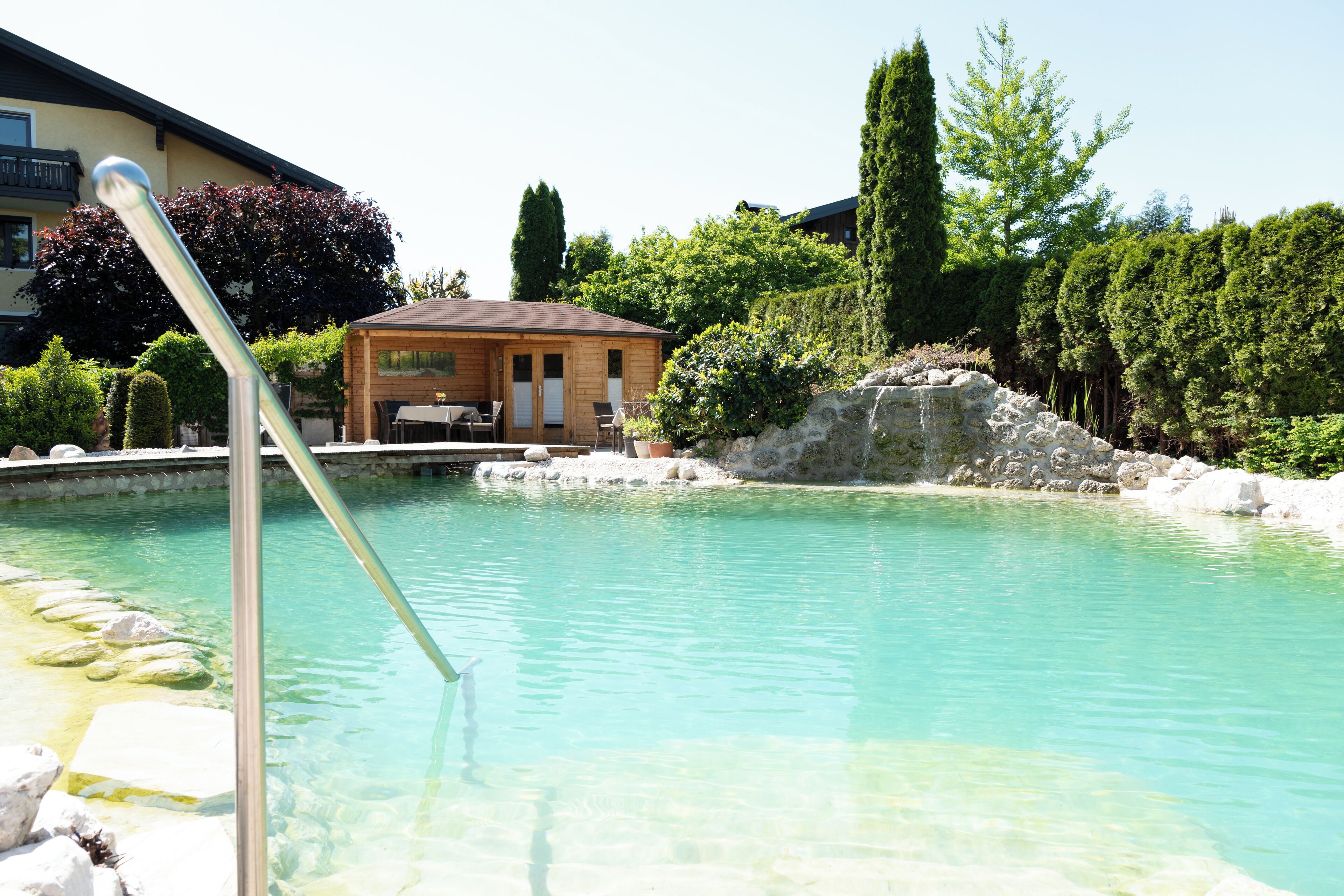 A clear pool with a wooden deck and a small building in the background.