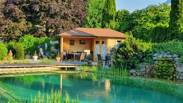 Un área de piscina al aire libre tranquila con una terraza de madera, asientos y un muro de piedra.