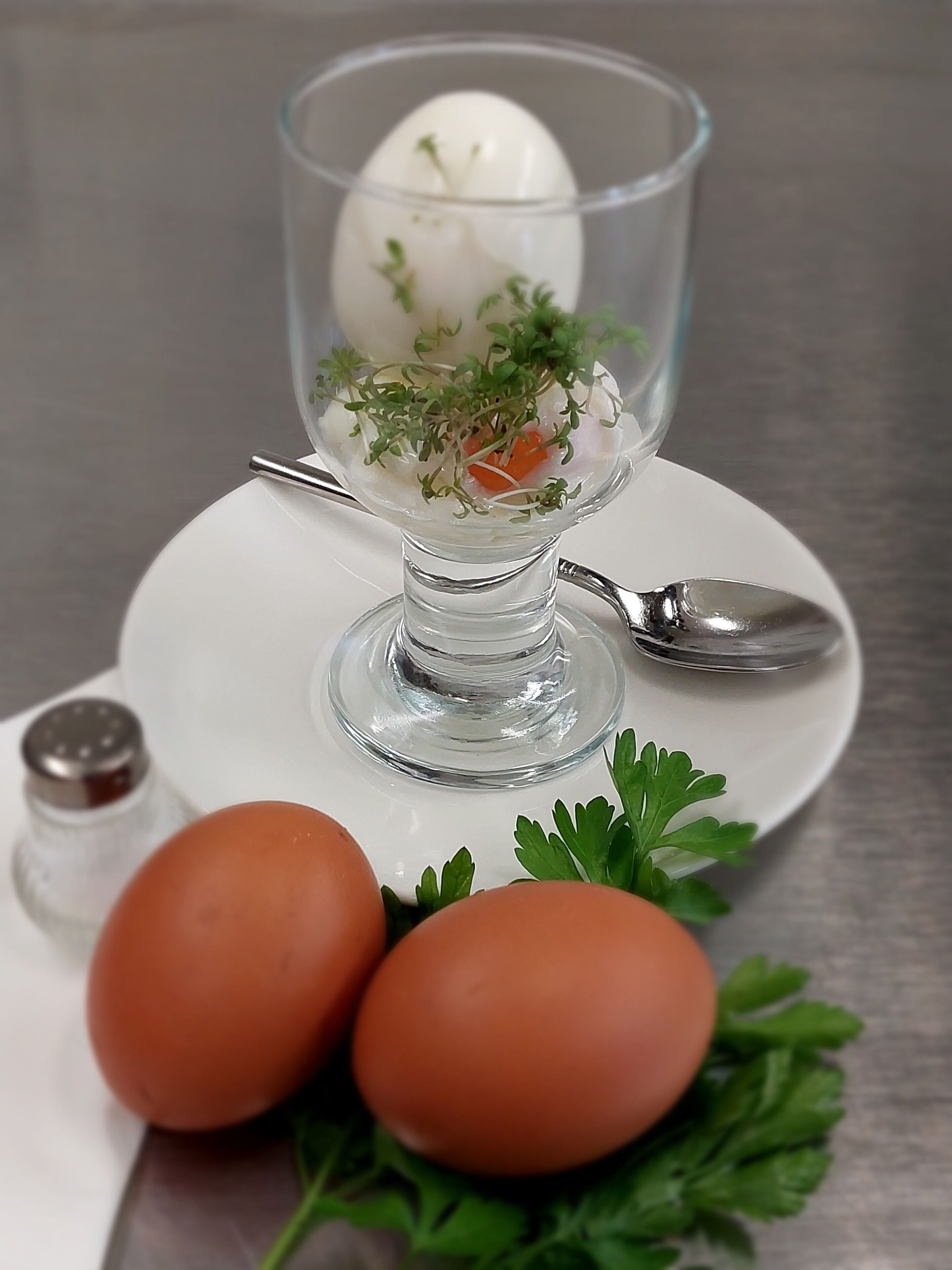 Un recipiente de vidrio transparente con un huevo blanco y hierbas verdes sobre un plato blanco, acompañado de dos huevos enteros y una ramita de perejil.