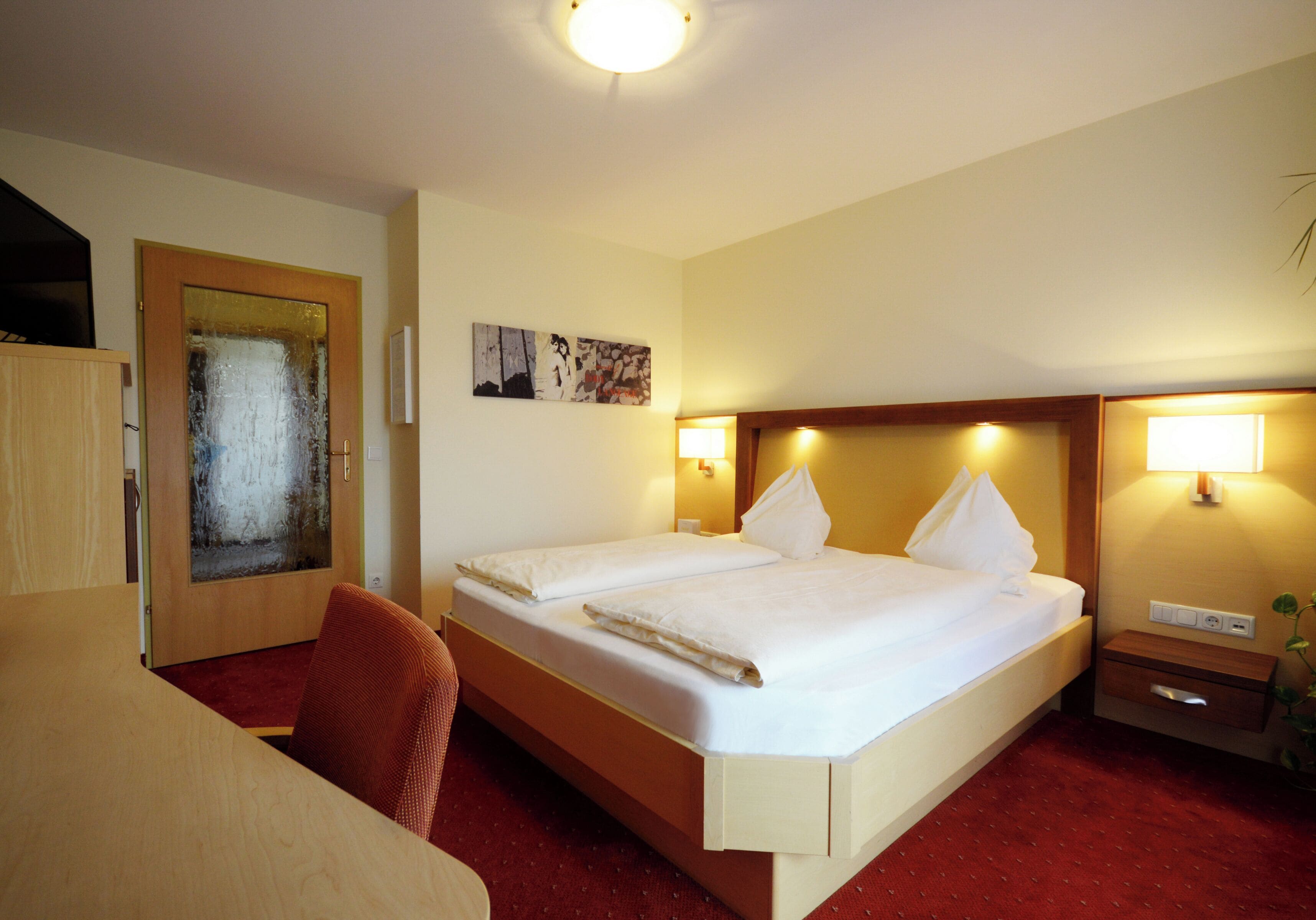 Habitación de hotel con una cama grande, un escritorio de madera y una silla. Hay dos lámparas de pared y un cuadro en la pared.