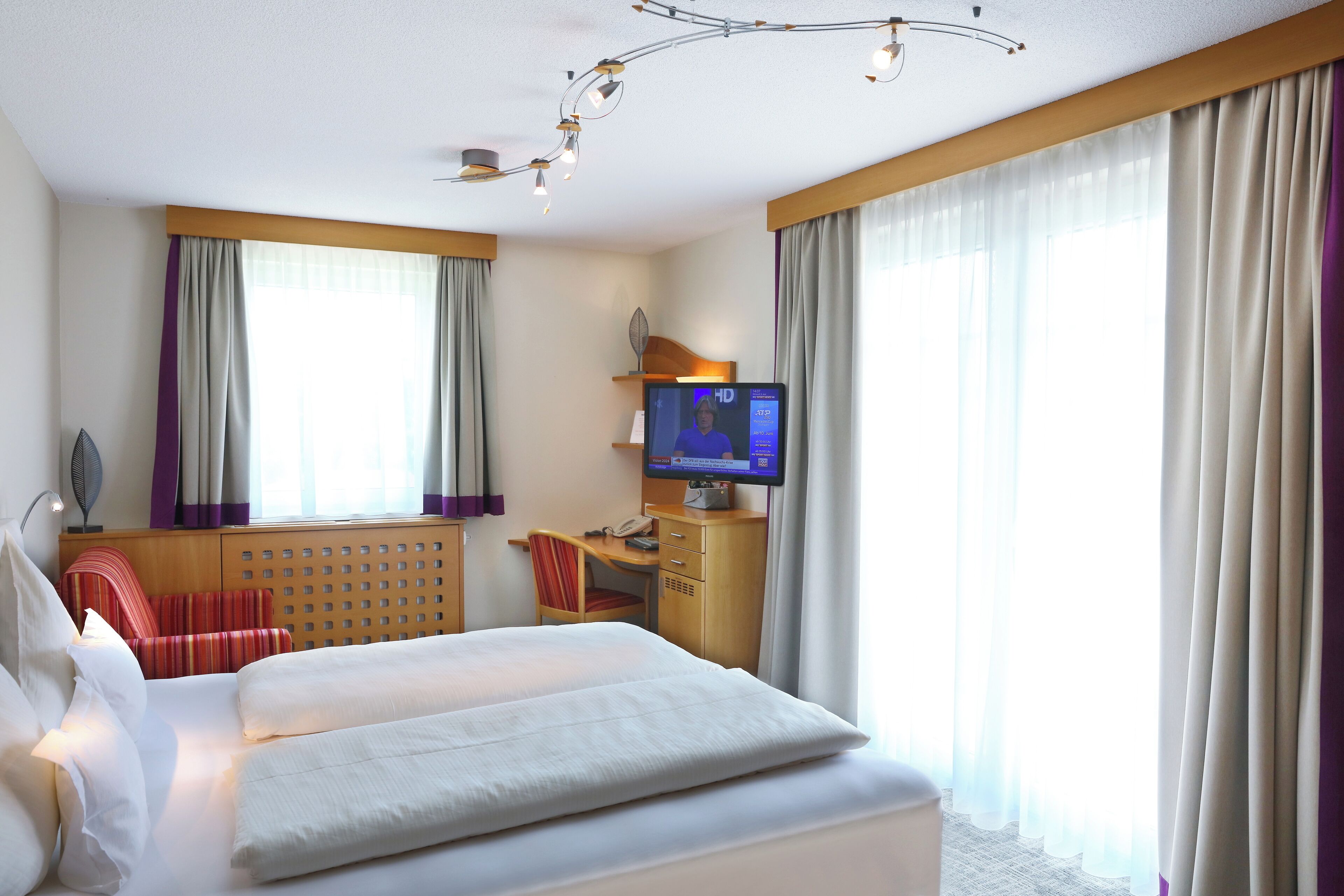A hotel room with a bed, a desk with a computer, a TV, and a window with curtains.