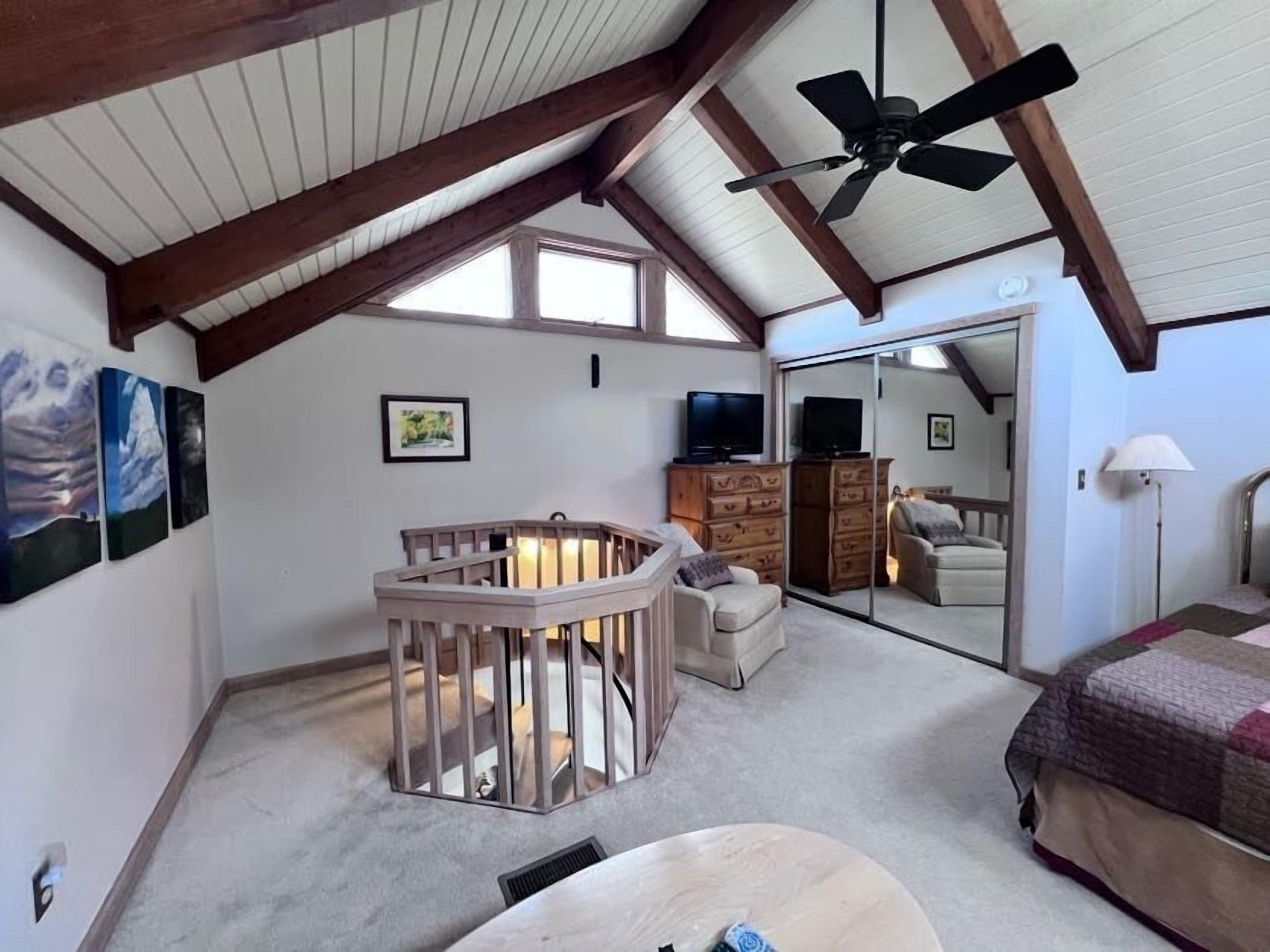 Een kamer met een hoge plafond, houten balken, een plafondventilator, een groot raam, een televisie, een bank, een stoel, een dressoir en een bed met quilt.