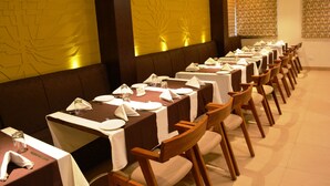 A long dining table with white tablecloths and wooden chairs, set up in a room with patterned wallpaper and a decorative wall panel.