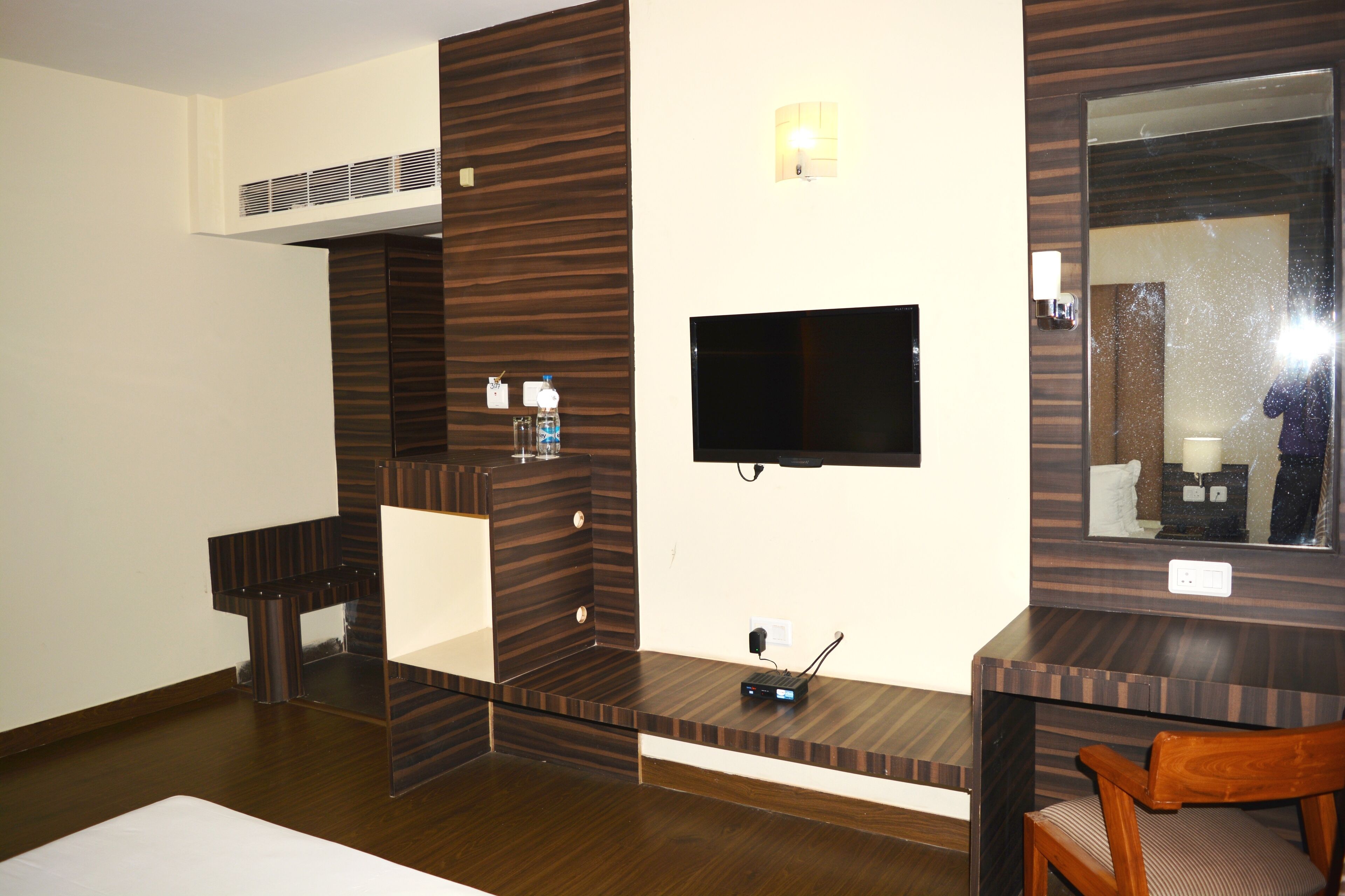 A hotel room with a flat-screen TV mounted on the wall, a wooden vanity with a mirror, and a small table with a chair.