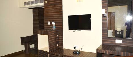 A hotel room with a flat-screen TV mounted on the wall, a wooden vanity with a mirror, and a small table with a chair.