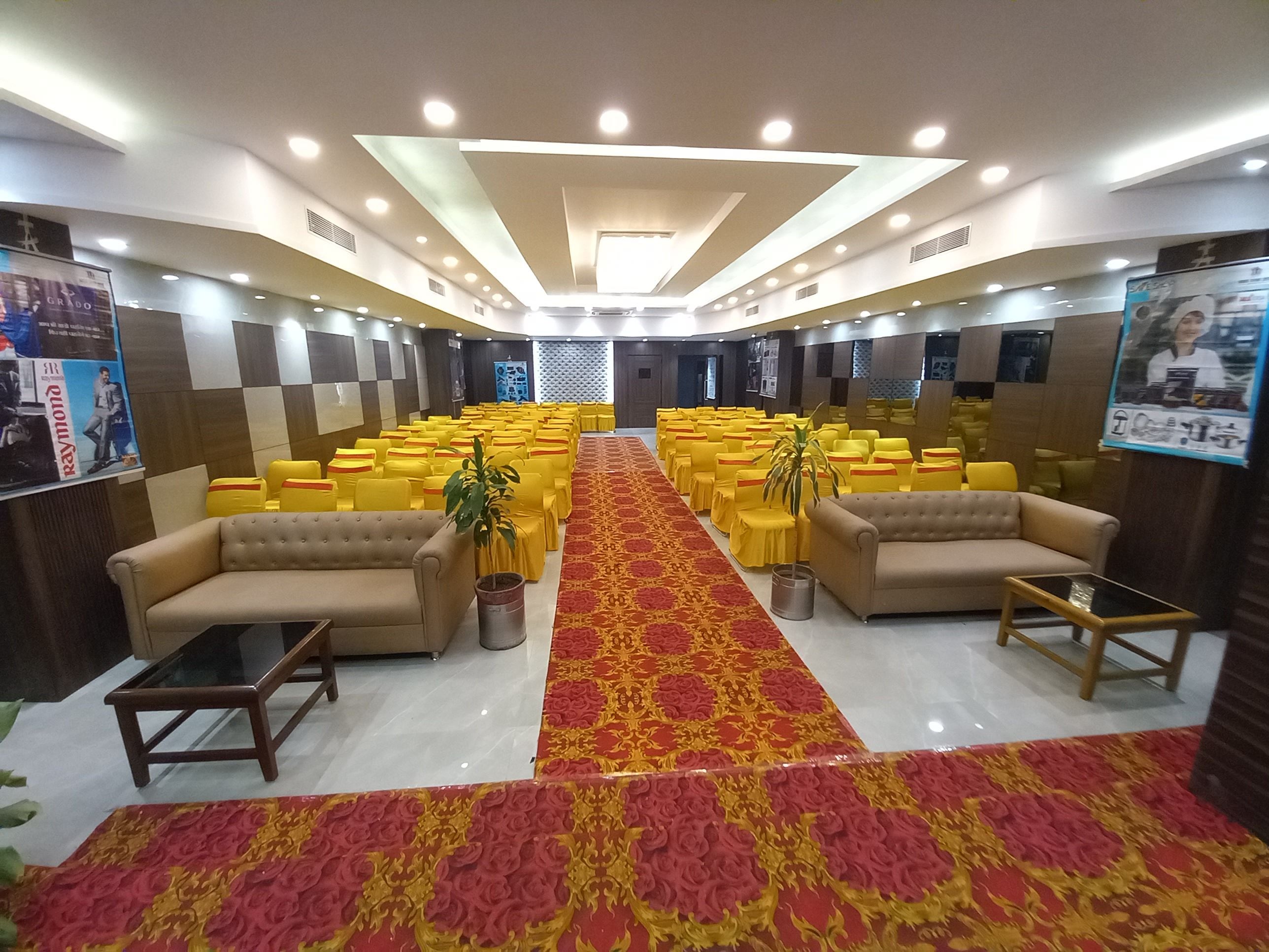 A conference room with rows of yellow chairs, two sofas, a patterned carpet, and a ceiling with recessed lighting.